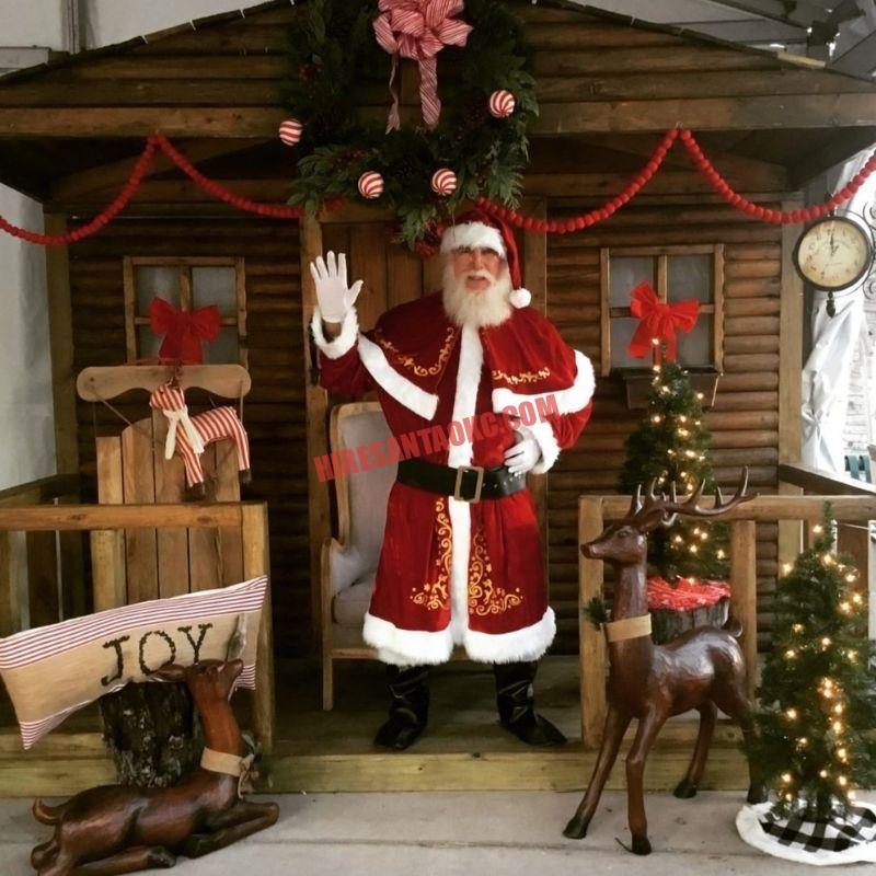 Real bearded Santa for hire in Oklahoma City waving from a log cabin porch beside a carved reindeer.