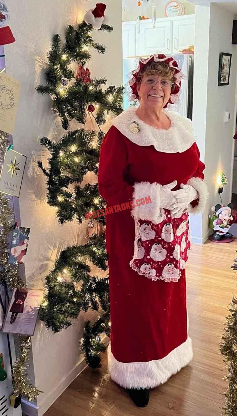 Mrs. Claus for hire in Oklahoma City standing beside a Christmas tree in a red dress.