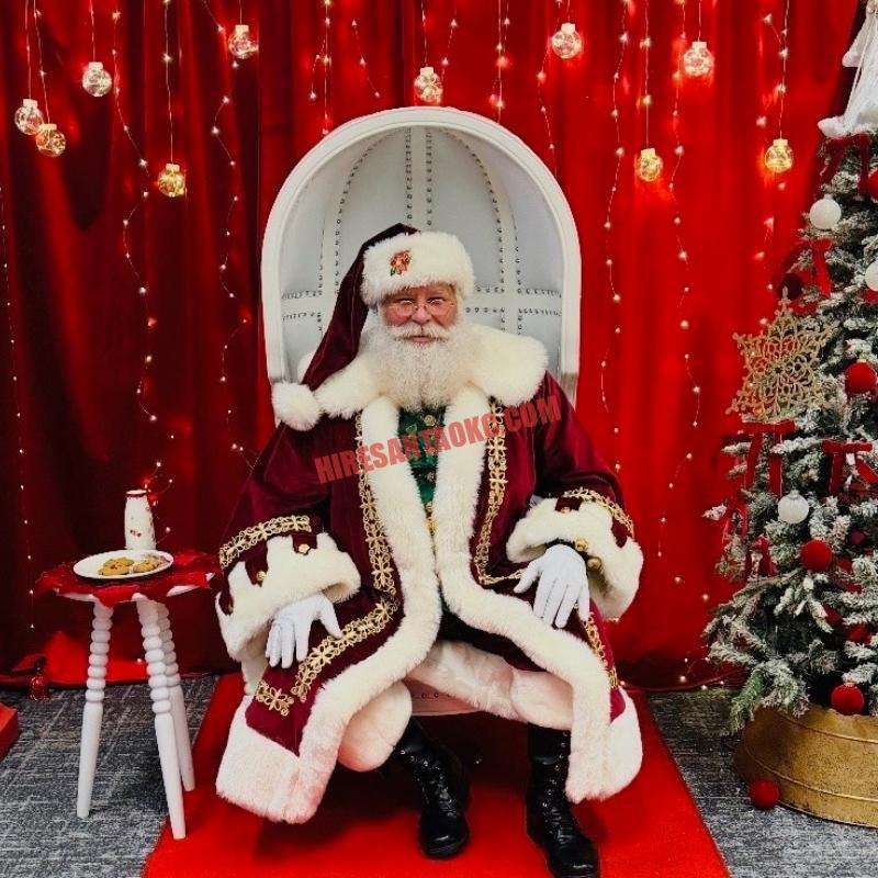 Santa for hire in Edmond, OK seated in a white chair with milk and cookies on a side table.